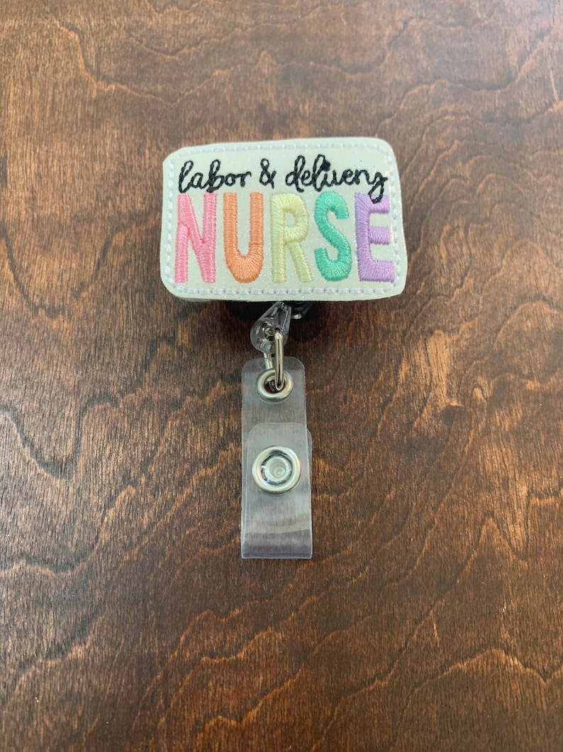 May include: A white badge holder with a colorful embroidered badge that says "labor & delivery NURSE". The badge is attached to a clear plastic retractable badge reel.