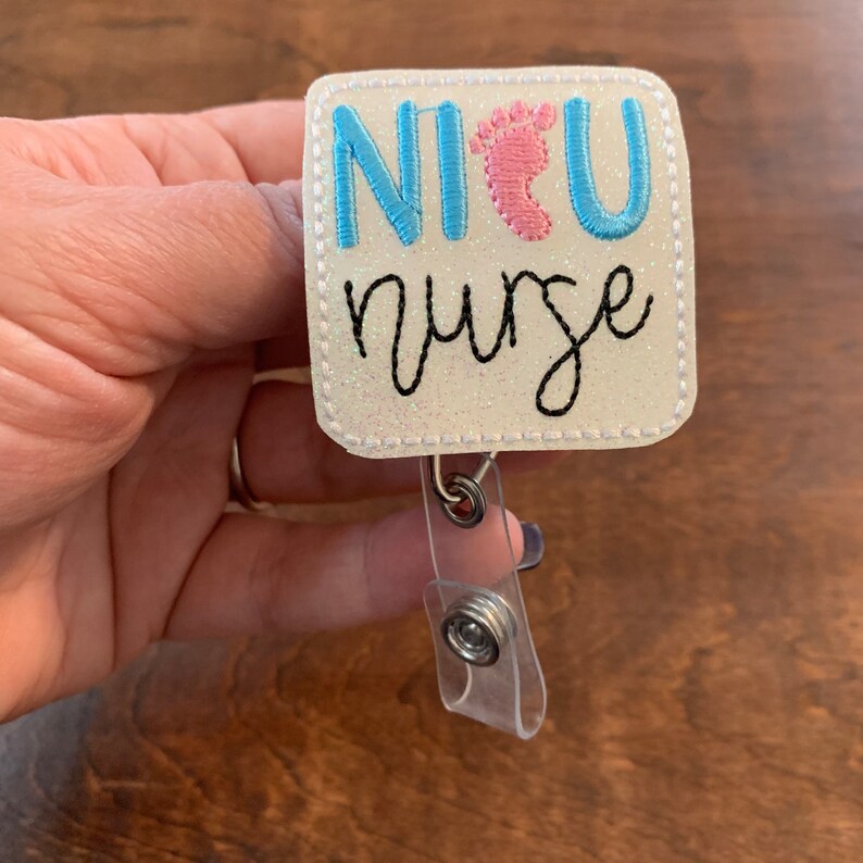 May include: A white, glittery badge reel with the text "NICU nurse" in blue and black script. The "U" is replaced with a pink baby footprint. The badge has a clear plastic strap and a metal clip.
