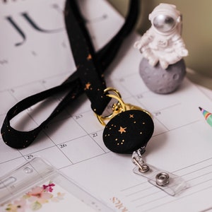 Lanyard and badge holder laying on a desk with a calendar. Fabric lanyard is black with metallic gold stars and gold hardware. A matching badge reel is hanging from lanyard. Clear ID Holder laying on desk.