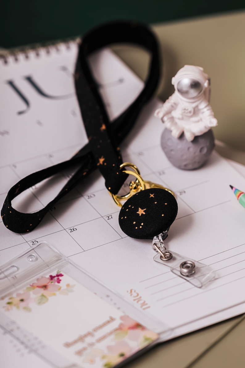 Lanyard and badge holder laying on a desk with a calendar. Fabric lanyard is black with metallic gold stars and gold hardware. A matching badge reel is hanging from lanyard. Clear ID Holder laying on desk.