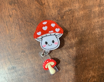 May include: A red and white mushroom with a smiling face, attached to a retractable badge holder with a smaller red and white mushroom charm.