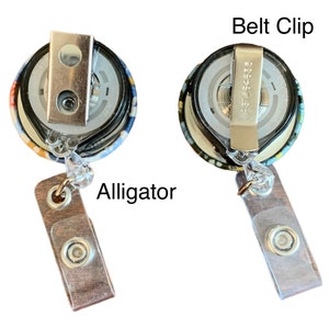 May include: A colourful fabric retractable badge holder with a metal alligator clip and a belt clip. The badge holder has a clear plastic strap and a brown wooden alligator clip.