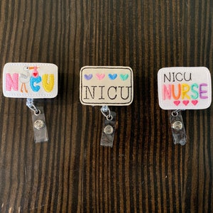 May include: Three white embroidered badge reels with colourful embroidery. The first reel says "NICU" with a stork and baby. The second reel says "NICU" with hearts. The third reel says "NICU NURSE" with hearts.
