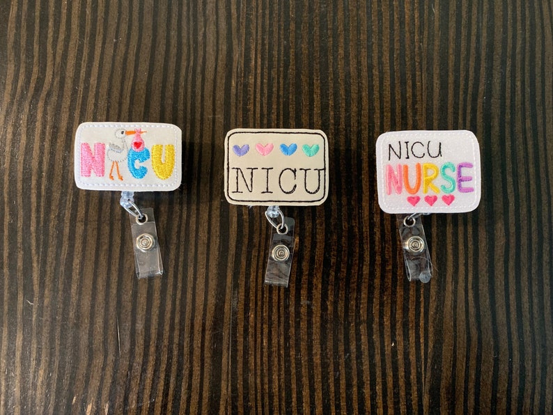 May include: Three white embroidered badge reels with colourful embroidery. The first reel says "NICU" with a stork and baby. The second reel says "NICU" with hearts. The third reel says "NICU NURSE" with hearts.