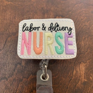 May include: A white badge holder with a glittery background and the text "labor & delivery NURSE" in rainbow colors. The badge holder has a clear plastic retractable badge reel.