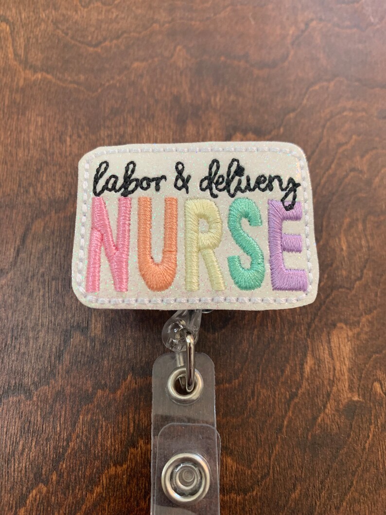 May include: A white badge holder with a glittery background and the text "labor & delivery NURSE" in rainbow colors. The badge holder has a clear plastic retractable badge reel.