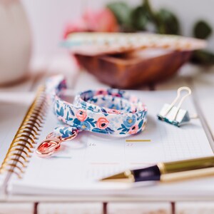 May include: A white and blue floral patterned lanyard with a gold key ring and a gold spiral bound notebook with a pen on top.