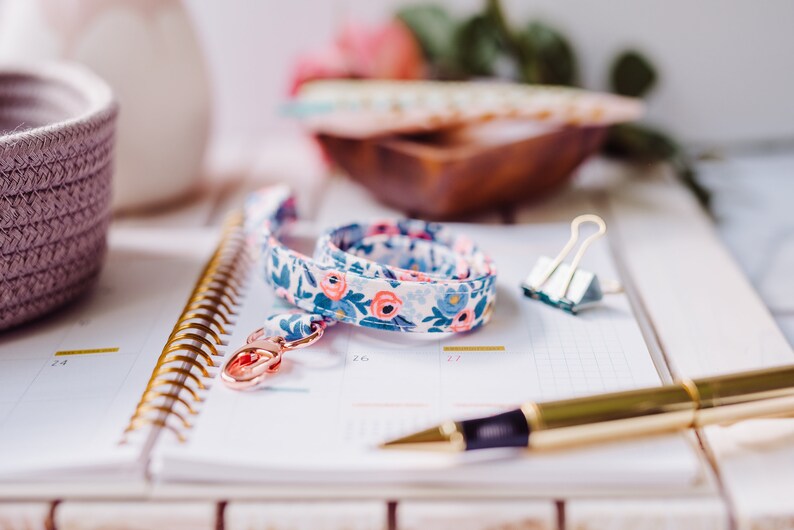 May include: A white and blue floral patterned lanyard with a gold key ring and a gold spiral bound notebook with a pen on top.