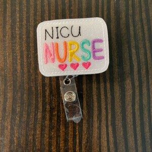 May include: White glitter badge holder with a rainbow embroidered "NICU NURSE" and three red hearts. The badge holder has a clear plastic retractable badge reel.