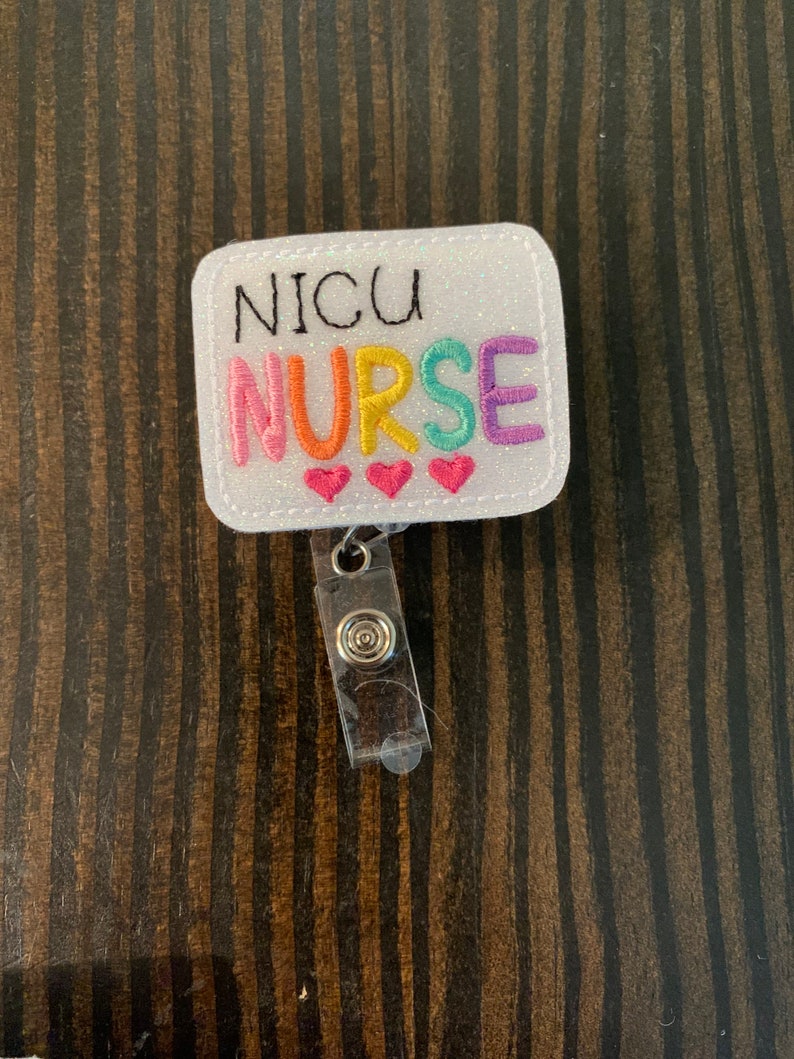 May include: White glitter badge holder with a rainbow embroidered "NICU NURSE" and three red hearts. The badge holder has a clear plastic retractable badge reel.