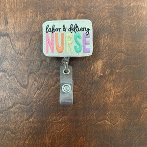 May include: A white rectangular badge with the text "labor & delivery NURSE" in rainbow colors. The badge is attached to a clear plastic retractable badge holder.