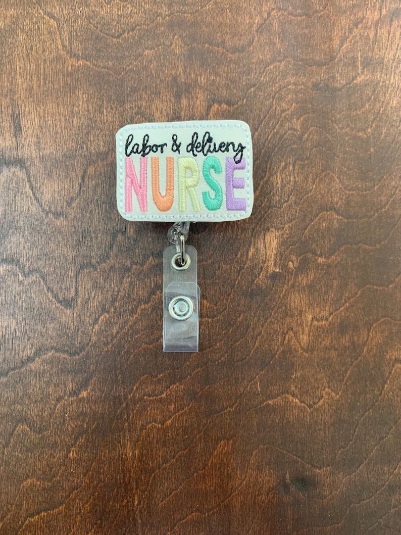 May include: A white rectangular badge with the text "labor & delivery NURSE" in rainbow colors. The badge is attached to a clear plastic retractable badge holder.