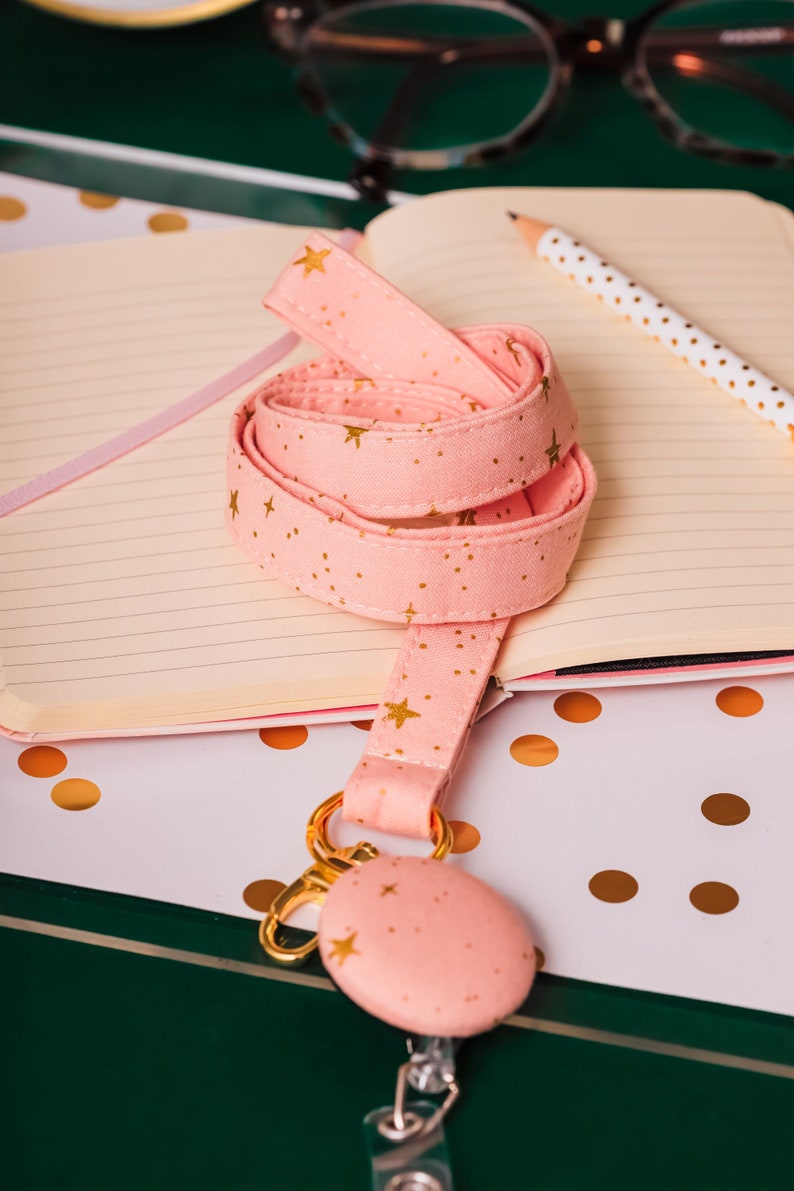 Soft pink fabric lanyard with metallic gold stars and gold hardware laying on a desk. A matching badge reel is hanging from the lanyard.