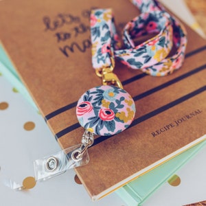 May include: A pink and blue floral patterned lanyard with a gold retractable badge holder and a matching floral button. The lanyard is on top of a stack of books, one of which is a recipe journal.