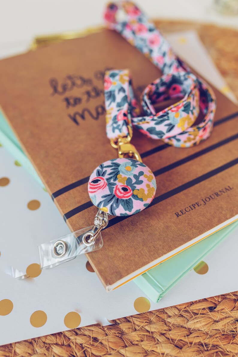 May include: A pink and blue floral patterned lanyard with a gold retractable badge holder and a matching floral button. The lanyard is on top of a stack of books, one of which is a recipe journal.