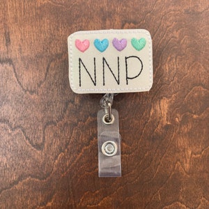 May include: White badge holder with a white rectangle with four pastel colored hearts and the text "NNP" in black. The badge holder has a clear plastic retractable badge reel.