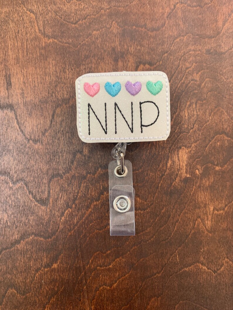 May include: White badge holder with a white rectangle with four pastel colored hearts and the text "NNP" in black. The badge holder has a clear plastic retractable badge reel.