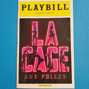 May include: Playbill for the musical "La Cage aux Folles" at the Longacre Theatre. The playbill features a pink neon sign with the title of the show and silhouettes of dancers in the background.