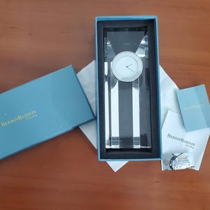 Può includere: Un orologio di cristallo Reed & Barton in una scatola blu. L'orologio ha un quadrante bianco e un corpo in cristallo rettangolare. La scatola è aperta, rivelando l'orologio e l'imballaggio. La scatola ha il logo Reed & Barton.