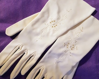 Könnte beinhalten: Ein Paar weiße lange Handschuhe mit zarter Blumenstickerei. Die Stickerei zeigt kleine weiße Blüten und Blätter mit goldenen Akzenten.