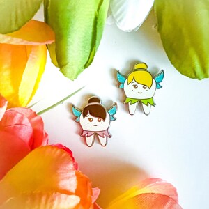 May include: Two enamel pins featuring cartoon fairies with wings and tooth-shaped bodies. The fairy on the left has purple hair and a pink dress. The fairy on the right has blonde hair and a green dress.