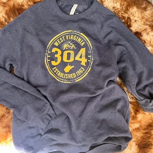 Puede incluir: Una sudadera azul marino con un gr&aacute;fico circular amarillo que dice "West Virginia 304 Established 1863". El gr&aacute;fico incluye una cordillera y la forma del estado. La sudadera est&aacute; hecha de un material suave.
