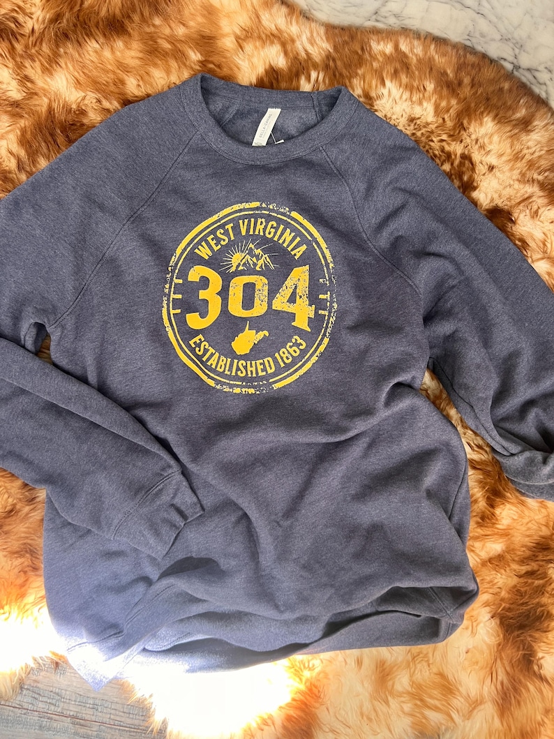 Puede incluir: Una sudadera azul marino con un gr&aacute;fico circular amarillo que dice "West Virginia 304 Established 1863". El gr&aacute;fico incluye una cordillera y la forma del estado. La sudadera est&aacute; hecha de un material suave.