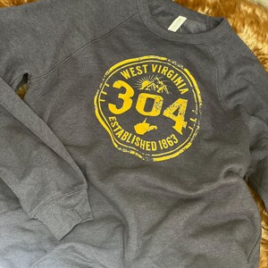 Puede incluir: Una sudadera gris oscuro con un gr&aacute;fico circular amarillo. El gr&aacute;fico dice "WEST VIRGINIA 304 ESTABLISHED 1863" con una monta&ntilde;a y el contorno del estado. La sudadera tiene mangas largas y cuello redondo.