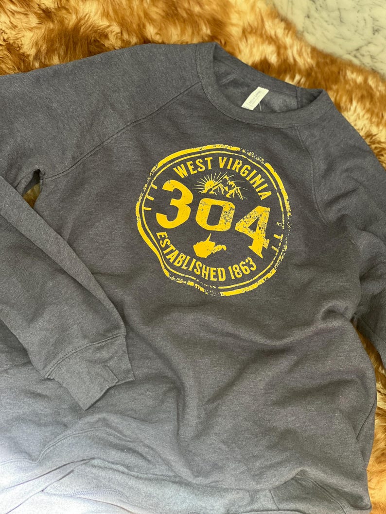 Puede incluir: Una sudadera gris oscuro con un gr&aacute;fico circular amarillo. El gr&aacute;fico dice "WEST VIRGINIA 304 ESTABLISHED 1863" con una monta&ntilde;a y el contorno del estado. La sudadera tiene mangas largas y cuello redondo.