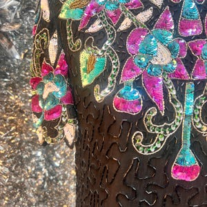 May include: Black sequined top with vibrant floral embroidery. The embroidery incorporates pink, teal, and gold sequins, with green and silver details. The top has short sleeves and a detailed, textured design.