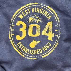 Puede incluir: Una sudadera azul marino con un gr&aacute;fico amarillo desgastado. El gr&aacute;fico es un dise&ntilde;o circular con las palabras "WEST VIRGINIA", "304" y "ESTABLISHED 1863". Un gr&aacute;fico de monta&ntilde;a y sol est&aacute; en el centro.