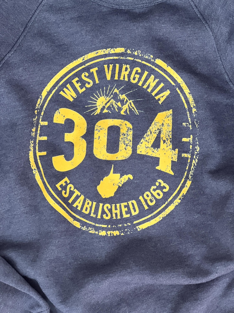 Puede incluir: Una sudadera azul marino con un gr&aacute;fico amarillo desgastado. El gr&aacute;fico es un dise&ntilde;o circular con las palabras "WEST VIRGINIA", "304" y "ESTABLISHED 1863". Un gr&aacute;fico de monta&ntilde;a y sol est&aacute; en el centro.