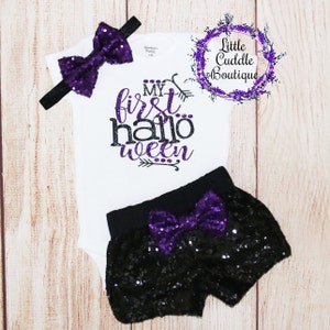 May include: A white baby bodysuit, black sequin shorts, and a headband with a purple sequin bow. The bodysuit has the text "My First Halloween" in purple and black. A matching purple bow is on the shorts.