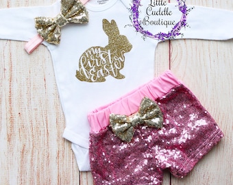 May include: A baby girl's Easter outfit with a white bodysuit featuring a gold glitter bunny and the text "my first easter", pink sequin shorts with a gold glitter bow, and a gold glitter headband with a bow.