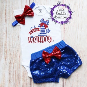 May include: A baby outfit set for a first birthday, featuring a white bodysuit with a red, white and blue glitter design that says "It's My 1st Birthday", a red sequin bow headband, and blue sequin shorts with a red sequin bow.