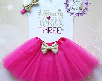May include: A white onesie with gold glitter lettering that says "Young Wild & Three" with a pink heart. The onesie is paired with a pink tulle tutu and a gold glitter bow.