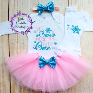 May include: A baby girl's first birthday outfit with a white bodysuit that says "It's Snow Much Fun to be One" in turquoise glitter lettering with silver glitter snowflakes. The outfit also includes a pink tulle tutu with a turquoise sequin bow, a white headband with a turquoise sequin bow, and white leg warmers with a turquoise snowflake.
