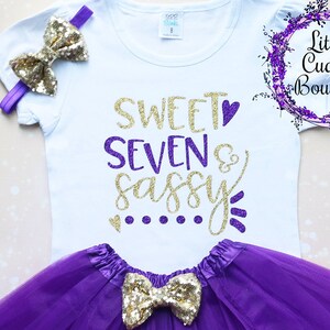 Puede incluir: Una camiseta blanca con el texto "Sweet Seven & Sassy" en purpurina dorada y morada. La camiseta se combina con una falda de tul morada y un lazo de purpurina dorada.