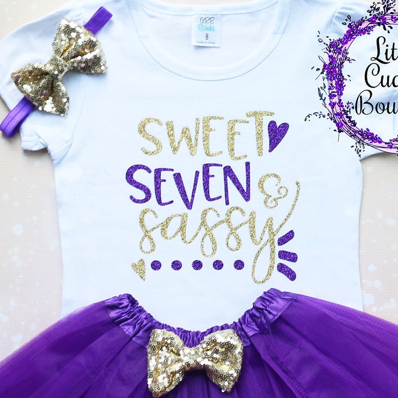 Puede incluir: Una camiseta blanca con el texto "Sweet Seven & Sassy" en purpurina dorada y morada. La camiseta se combina con una falda de tul morada y un lazo de purpurina dorada.