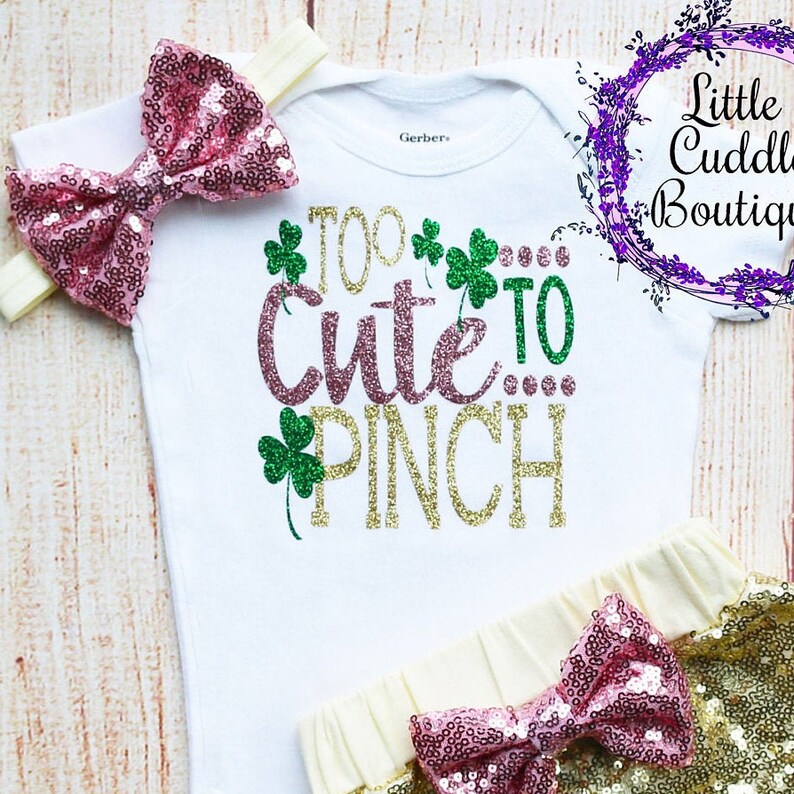 Pode incluir: Um macac&atilde;o branco para beb&ecirc; com o texto "Too Cute To Pinch" em glitter dourado e rosa. O macac&atilde;o &eacute; combinado com uma saia de lantejoulas douradas e uma faixa de cabelo com la&ccedil;o de lantejoulas rosa.