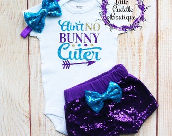 May include: A white baby onesie with the text "Ain't No Bunny Cuter" in gold and blue glitter. The onesie is paired with purple sequined shorts with a blue bow and a blue headband with a blue bow.