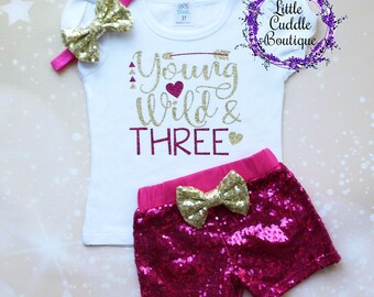 May include: A white t-shirt with gold glitter text that says "Young Wild & Three" with a pink heart and arrow. The shirt is paired with pink sequined shorts with a gold bow.