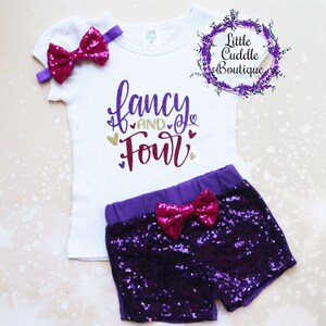 May include: A white shirt with the text "fancy and four" in purple and gold glitter. The shirt is paired with purple sequined shorts and a pink bow headband.