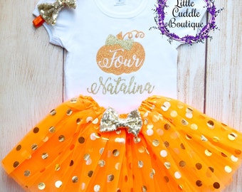 May include: Orange tulle skirt with gold polka dots and a gold bow. White t-shirt with a gold glitter pumpkin and the text "four" and "Natalina".