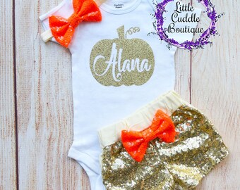 May include: A white onesie with a gold glitter pumpkin and the name "Alana" on it. The onesie is paired with gold sequin shorts with an orange bow. The outfit is perfect for a baby girl's first Halloween.