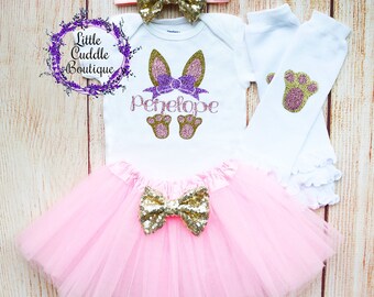 May include: A pink tulle tutu with a gold glitter bow, a white bodysuit with a pink glitter bunny and the name Penelope, a gold glitter headband with a bow, and white leg warmers with pink glitter paw prints.