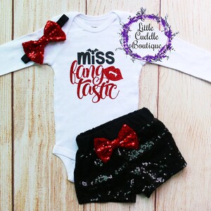 May include: A white long-sleeved bodysuit with the text "miss fangtastic" in red glitter. The bodysuit has a red glitter bow on the left sleeve. A pair of black sequined shorts with a red glitter bow.