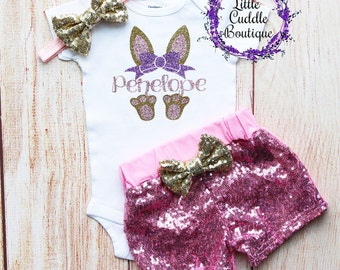 May include: A white baby bodysuit with a bunny ear design and the name "Penelope" in glitter. The outfit includes pink sequin shorts, a matching headband, and gold bow accents. The words "Little Cuddle Boutique" are also visible.