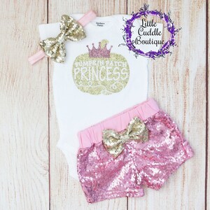 Peut inclure: Un body blanc avec un lettrage pailleté doré qui dit "Pumpkin Patch Princess" et un nœud pailleté doré. Le body est associé à un short pailleté rose avec un nœud pailleté doré.