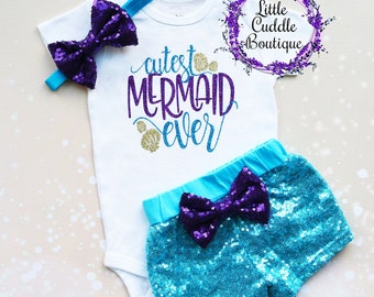 Peut inclure: Un body blanc avec le texte "cutest mermaid ever" en paillettes dorées et lettres violettes. Le body est associé à un short en sequins turquoise et un nœud en sequins violets. Un bandeau turquoise avec un nœud en sequins violets est également inclus.